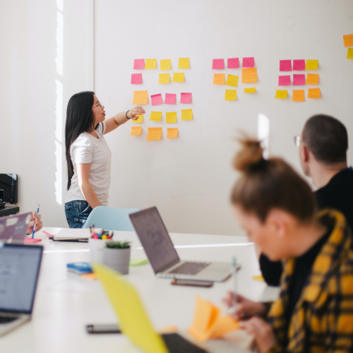 A woman gives a presentation, pointing to sticky notes on the wall, while her co-workers watch and take notes on their laptops.